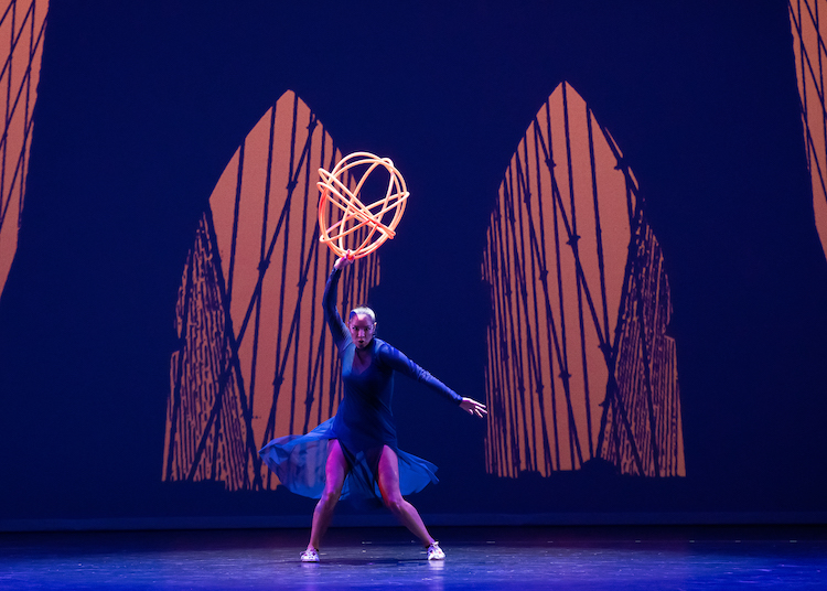 native American hoop dancer dressed in bright blue with a ball of interlocking hoops over her head, dances center stage against the background of a projected Brooklyn Bridge