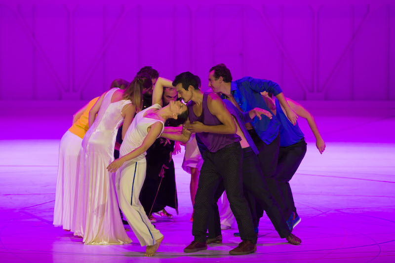 A group of dancers and opera singers huddle in a circle behind a magenta cyclorama background.