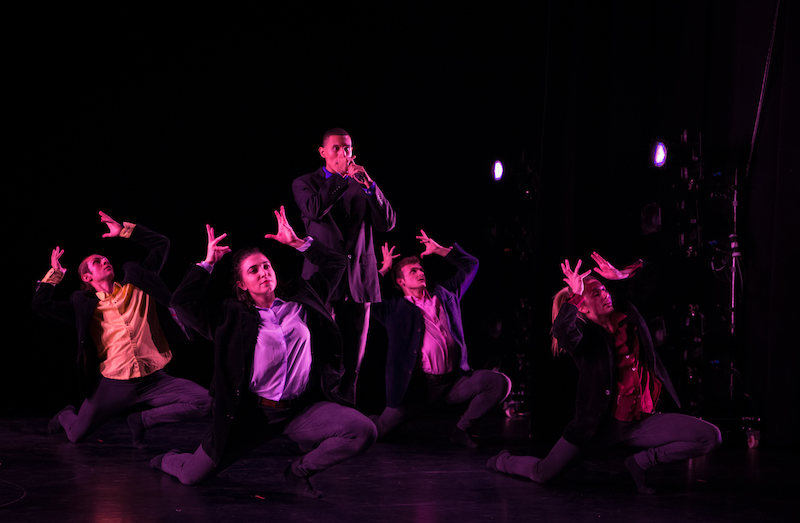 Dancers in black suits and button ups squat to the floor and their arms gesture above their heads as if they were placing a crown on top of their heads. One dancer stands with his hands in front of his mouth.
