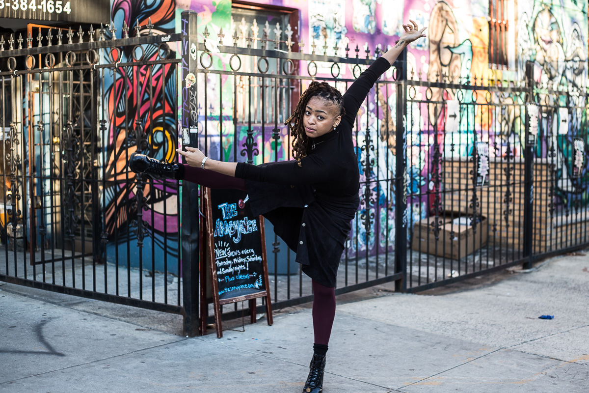A woman tilts her body, extending one leg and both arms, in front of an iron fence and colorful graffiti