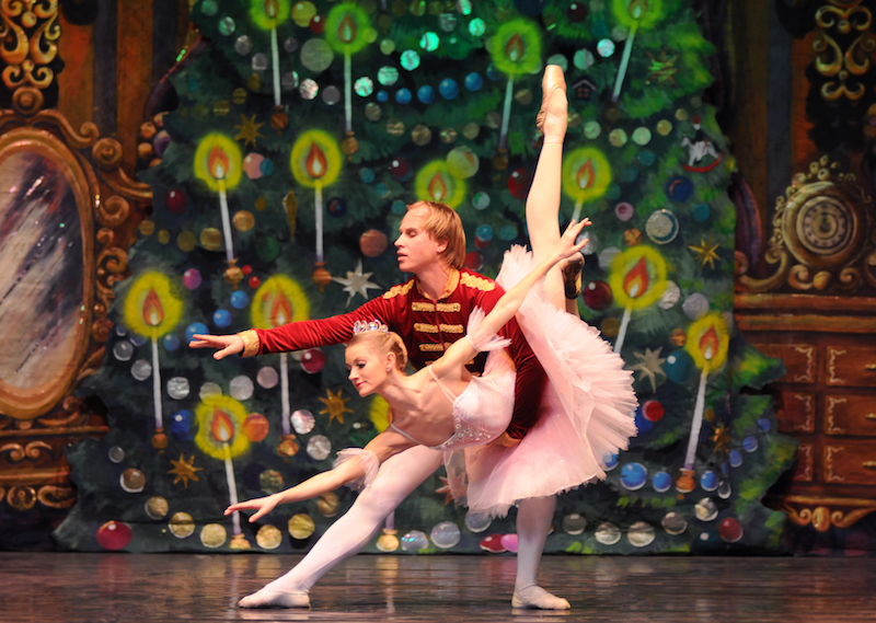 Russian ballet dancers pose in front of the Land of Sweets Nutcracker backdrop. The male in the role of prince holds his female partner in the dipped 