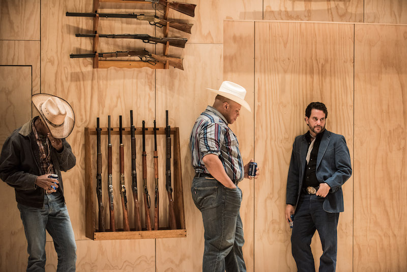 Three men stand against a wood-paneled wall. Two wear Cowboy hats. Rifles are affixed to the wall behind them.