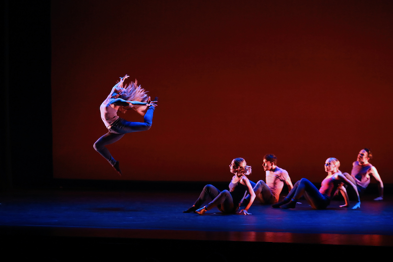 Four dancers sit and watch another dancer jump high into the air. Both of her legs are bent and her head tilts to the ceiling. Her blonde hair cascades around her.