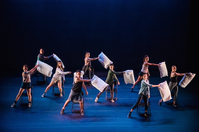The ensemble in street clothees each hold a pillow in their right hands and stand in front of a folding chair