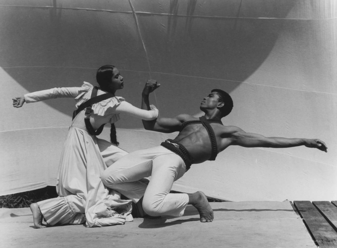 A woman in prairie dress links arms with a bare-chested man in a low backward lunge.