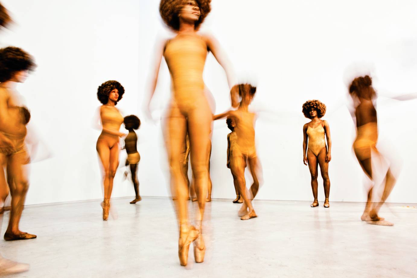 A roomful of women and men dancers in gold leotards. Some dancers are barelegged and others in shorts. Some wear pointe shoes and others are barefoot. All wear full Afro wigs.