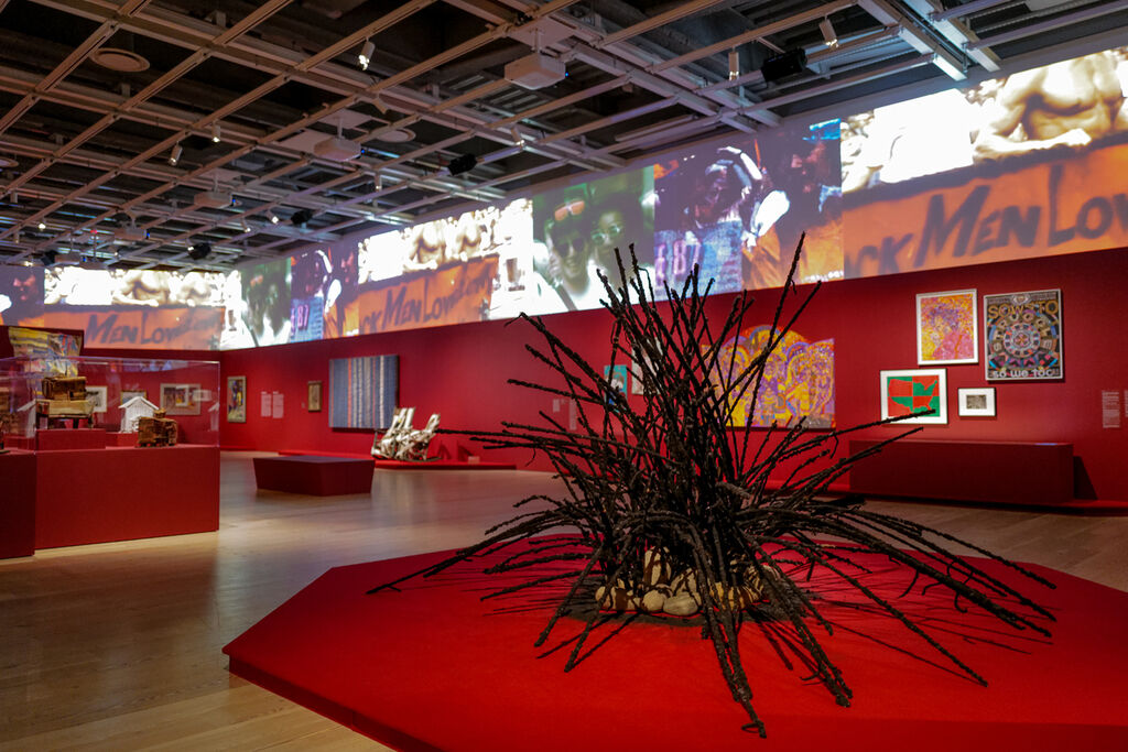 A view of the Whitney Museum exhibition space with the 6' high strip of video above the viewer. Exhibition paintings hung on red walls are situated behind a large spikey sculpture of black textured hair.