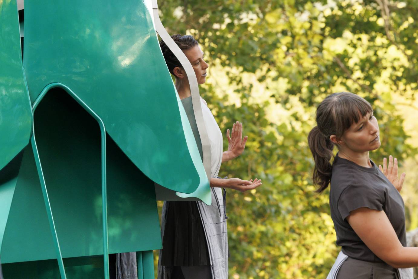 Two women dancers in gray and white tops lean close to a monumental green metal sculpture.