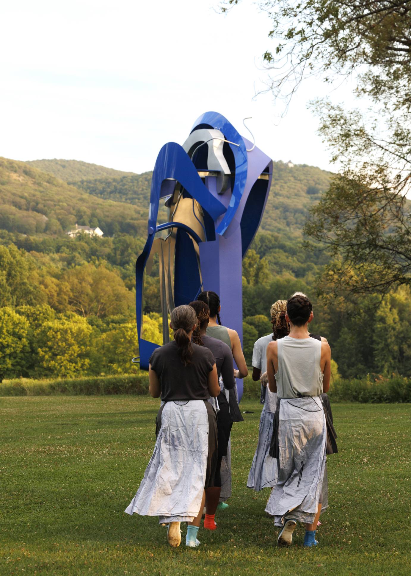 Six dancers two-by-two, their backs to us, approach a giant blue metal sculpture.