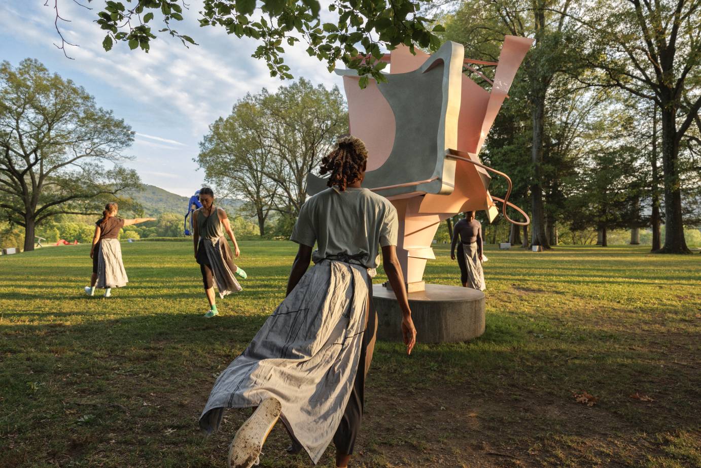 Four dancers flank a large pink metal sculpture situated in the grass with trees and hills in the background.