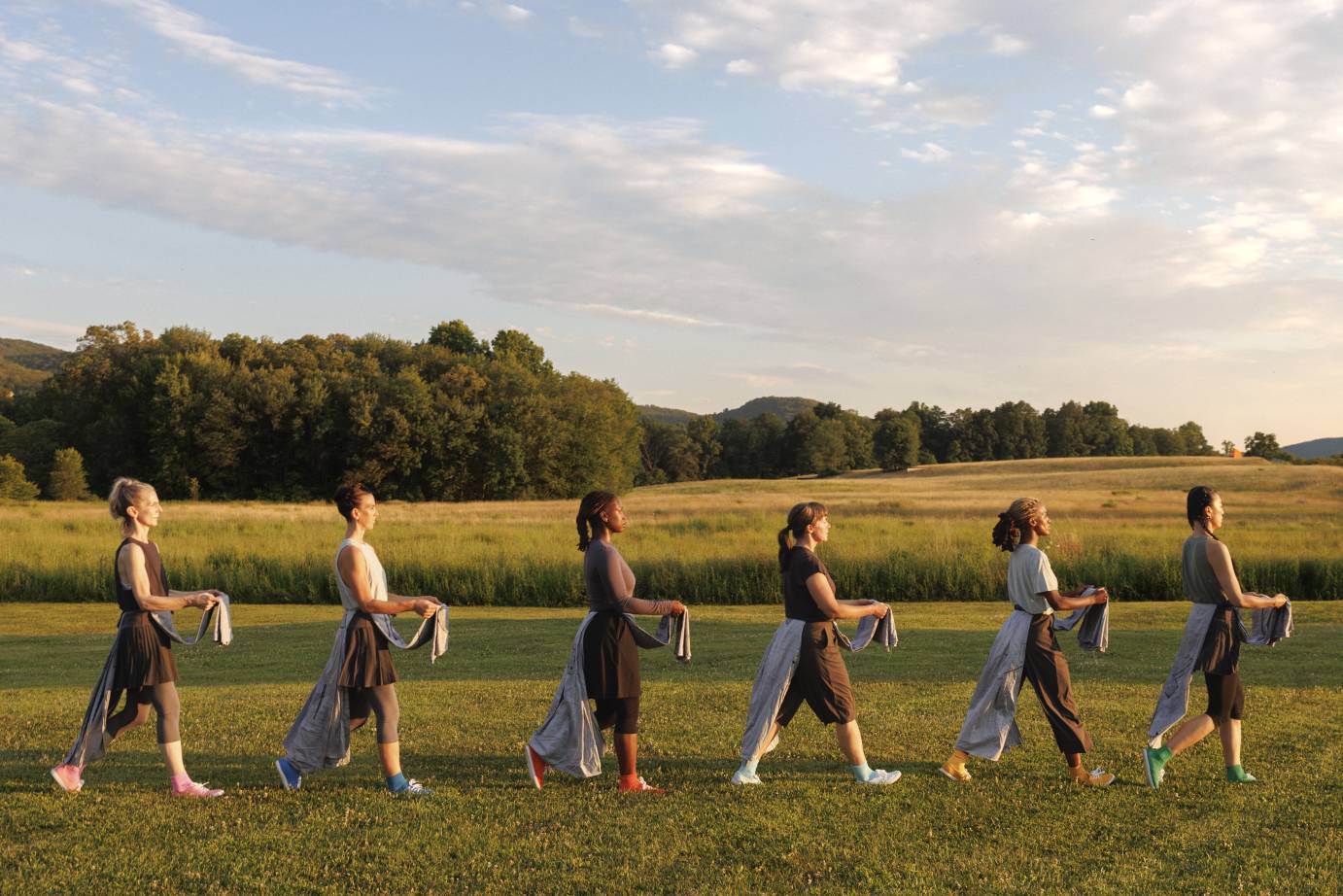 Six women dancers holding their folded aprons in front of their bodies walks in profile single file across the landscape