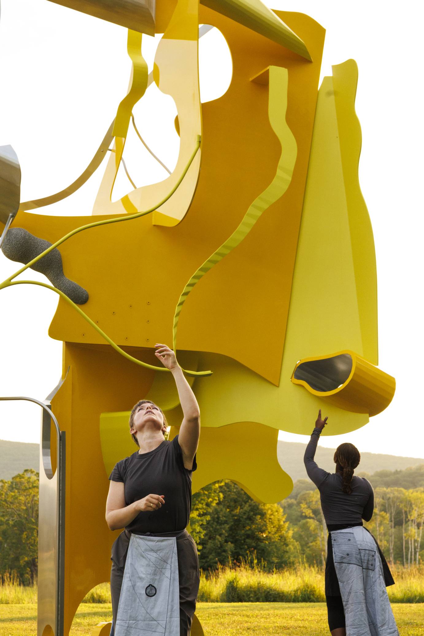 Two women dancers reach upward against the backdrop of a giant yellow metal sculpture with green hills in the background.