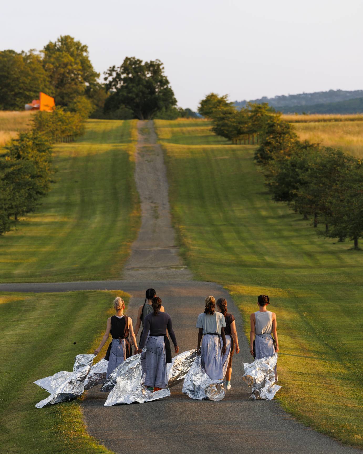 Six women dancers trailing silver blankets of fabric as they walk down a long gravel road into the distance. A giant ornage sculpture awaits.