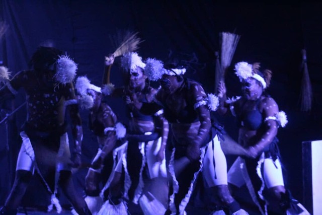 agroup of dancers with wave raffia strands in their fists dance in a processional they are wearing balck and white with white headresses and white dots painted on their bodies