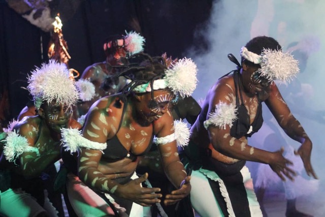 a group of women wearing black bikini like outfits andorned with white fringe hats and arm pieces dance joyously 