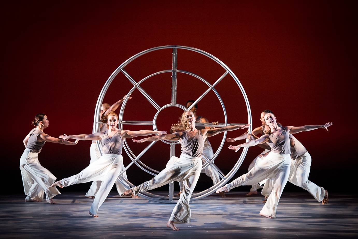 a company of dancers in white and grey maniputlate a gigantic silver wheel while three of their peers joyfully dance in front of them