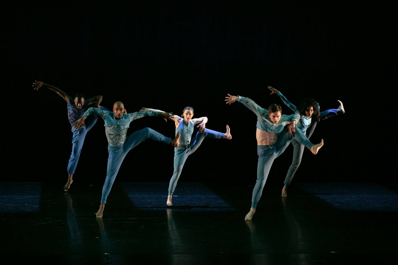 a group of five dancers dressed in blue leggings and blue toned tops stare at us as they perform an energetic kick to their sides with a bent leg...one arm grabs the extended leg or almost does as the other arm flies to its side. 