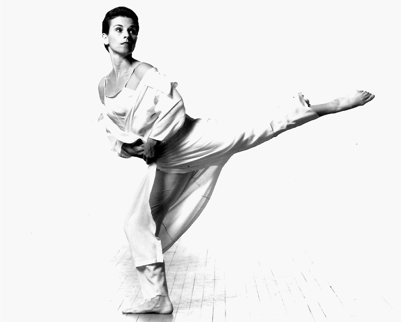 carolyn dorfman dressed in a white silky pant suit and jacket strikes an arabesque against a white background. she looks to her foot as if being called by someone