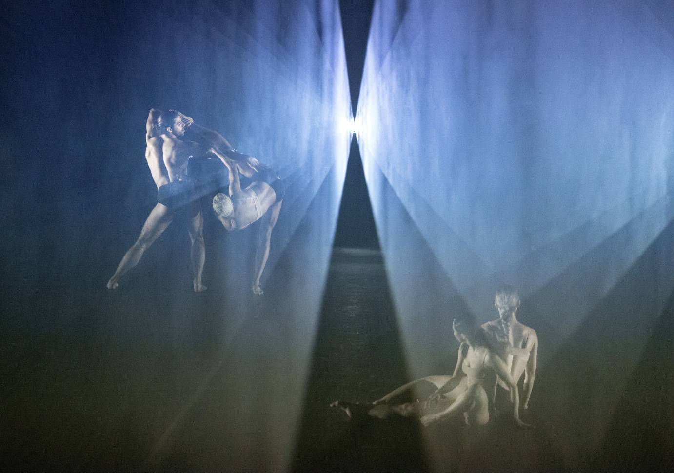 Two pairs of dancers behind a white lit scrim against a black background.