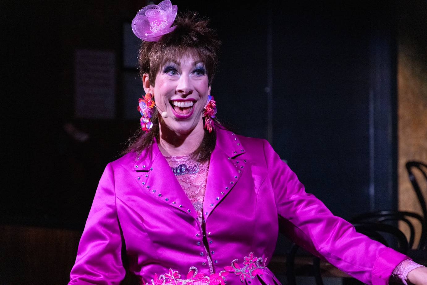 A short-haired woman with a bright smile and magenta flower near the top of her head, is dressed in a magenta jacket with her left arm extended