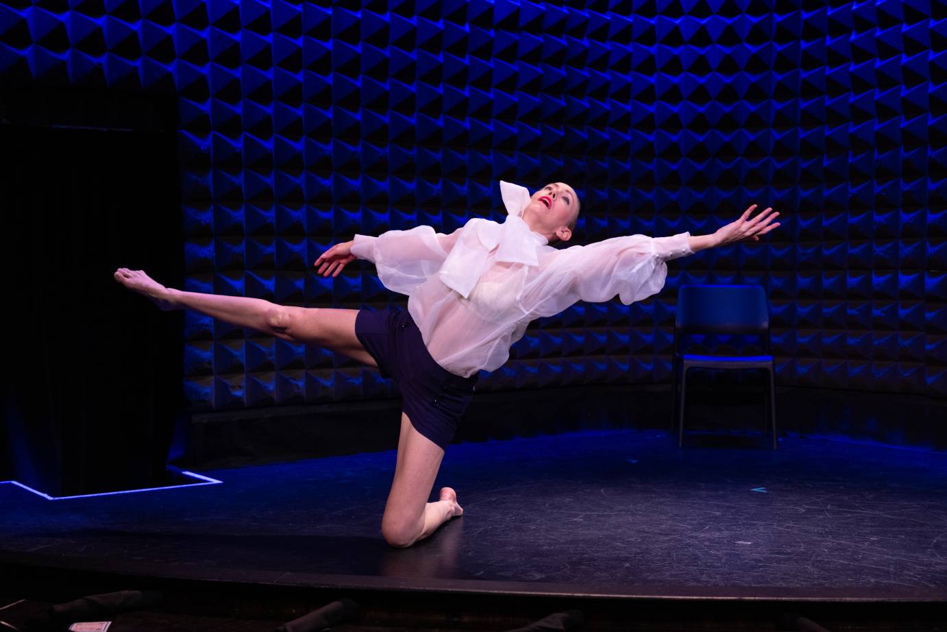 Woman in white billowy blouse and black shorts stretches to her left, left knee on floor while right leg lengthens. Her head is tilted to the ceiling.