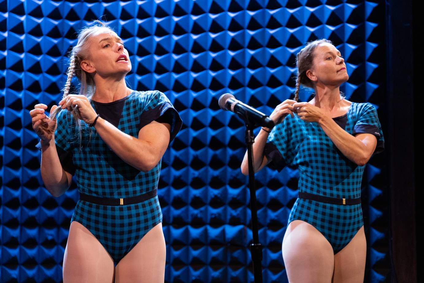 Two women with long hair, one blonde, the other brown, dressed in checked torquoise and black leotards braid their hair.