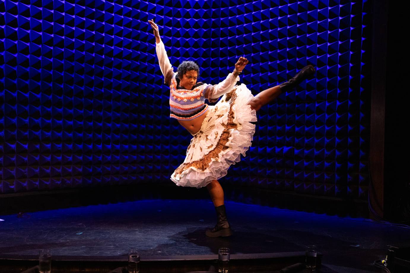 Woman dressed in a long sleeved striped sweater and big mid-length skirt with petticoats beneath in an arabeque, left leg high behind her head and both arms flung skyward