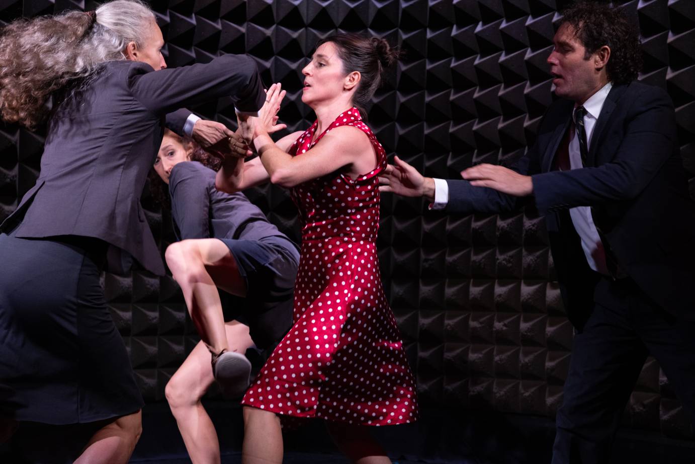 Four dancers intertwined with fraught expressesions - three (women and men) in business suits and a woman in a short sleeved white polka dot on red dress