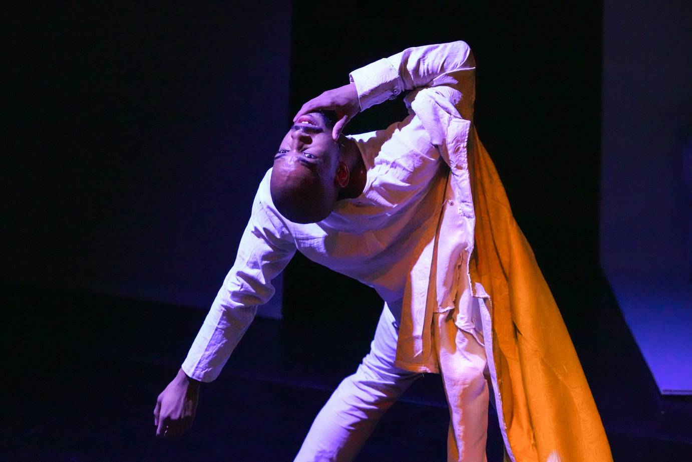 black man in white with a yellow lined jacket bends back towards the audience 