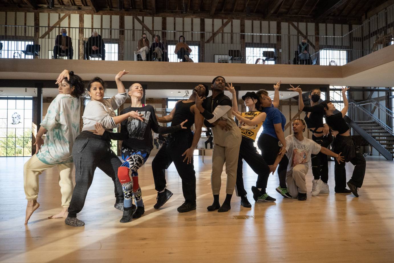 a  diagonal line of posing dancers, standing in work clothes looking like statues posing for a picture as they rehearse in studio Omari Wiles and Ballet Afrik