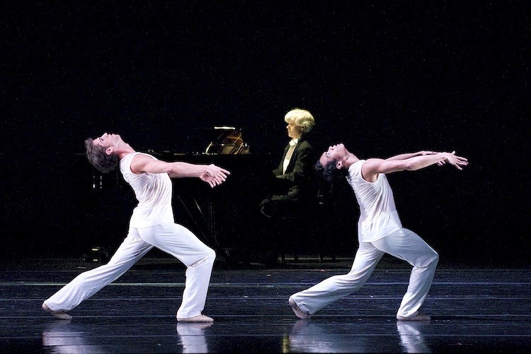 two men in white jump suits stand in a  wide forward lunge, as they arch their torsos back and extend their arms forward, behind them a whilte haired man plays a grand piano 