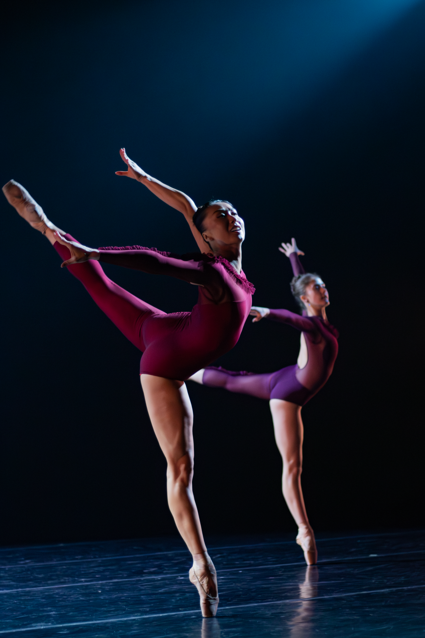 against a black background two female dancers hold a beautifully extended high arabesque... they are the perfect examples of beautiul muscular technique, gorgeous form 