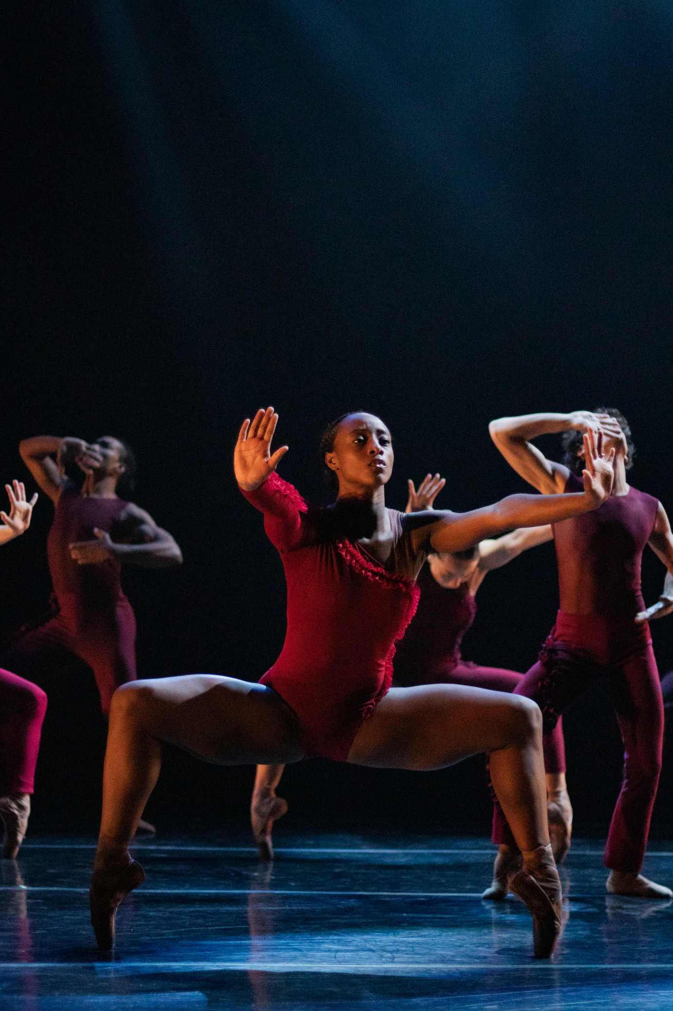 Black woman dancer in a red off the shoulder leotard holds a deep knee bend on pointe while staring out at the audience