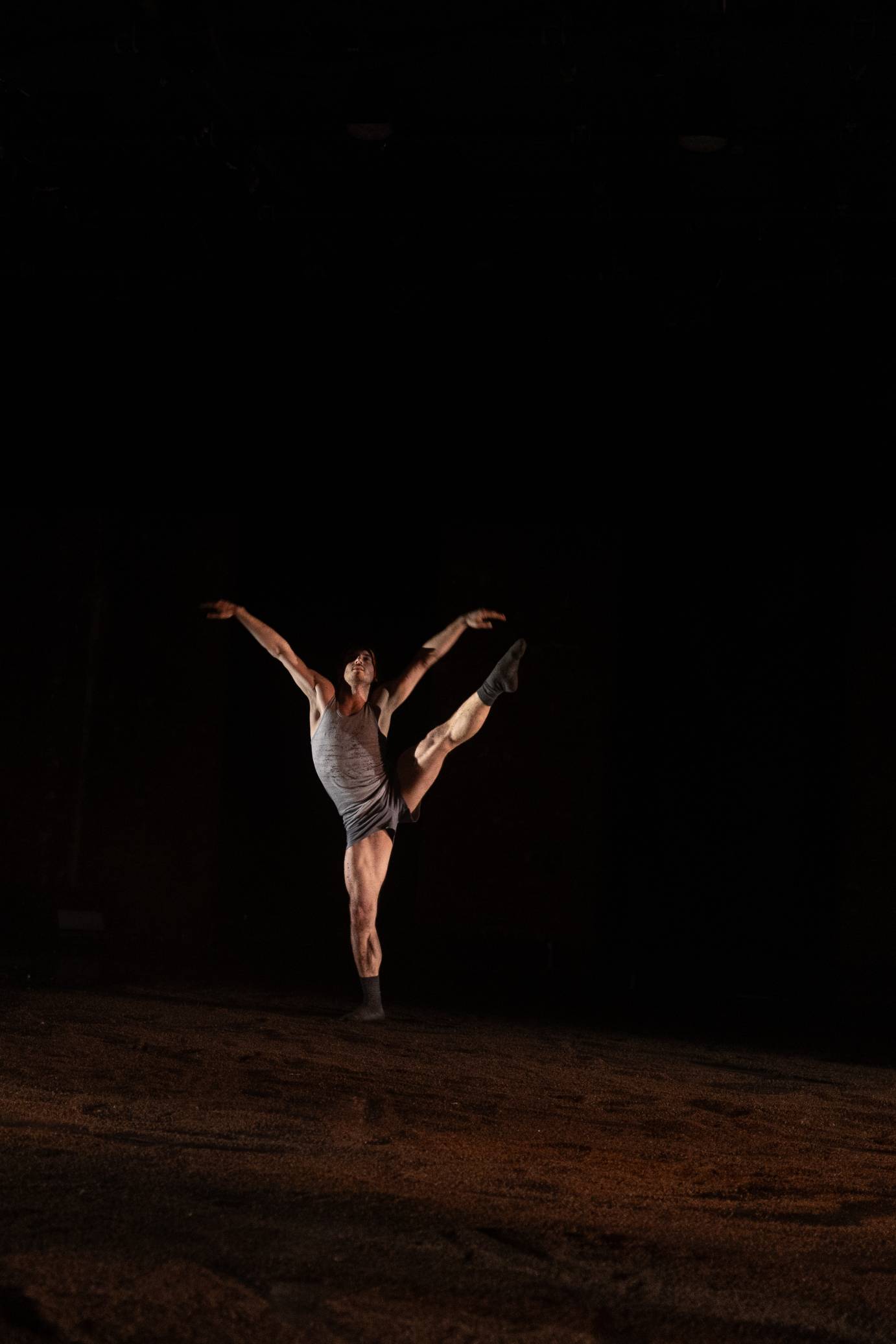 a man in dark grey shorts and a lighter gray top stands on one leg his other leg extended to the side, and his arms in a v shape, there is a sense of resignation in this pose...