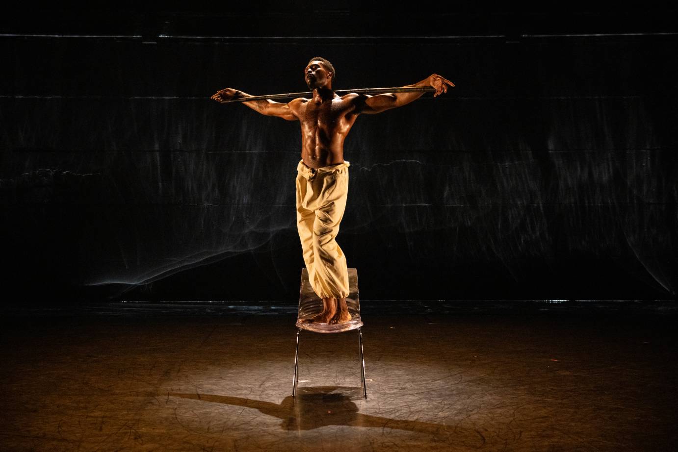 a Black man shining with sweat holds a stick across his shoulders as he balances on a chair. He looks like Christ on the Cross.