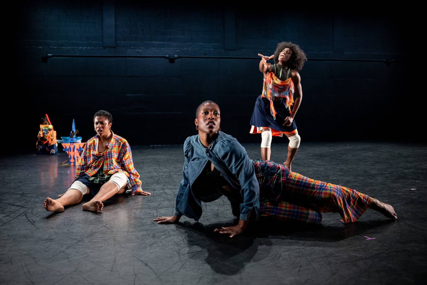 Three Black women form a triangle on stage. They are wearing colorful everyday clothes and each seems to be in their own world of communication... 