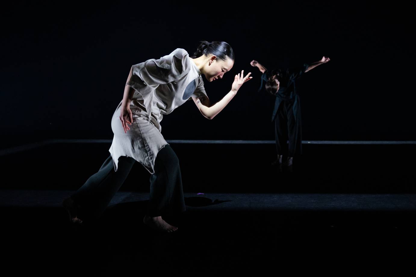 two figures on a dimly lit stage with tunics and loose pants the woman in the foreground is more brightly lit and we look at her profile, the woman in the back is bowing towards us. She blends into the black background and floor save for her arms, hair and parts of her face.