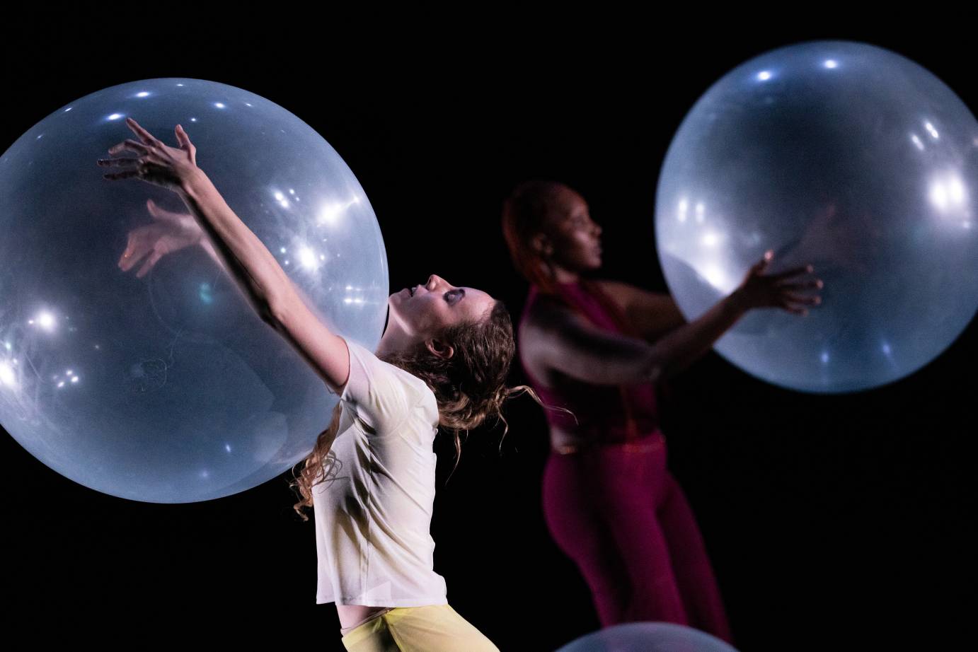 two dancers face away from each other holding  huge clear balloons