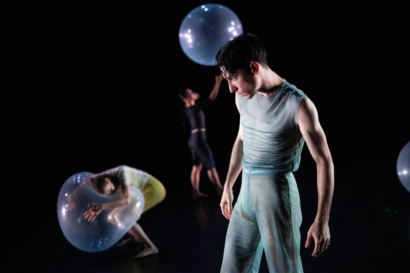 dancers in the background of this scene play with balloons  as  a male presenting figure in sheer green body hugging fabric looks on pensively 