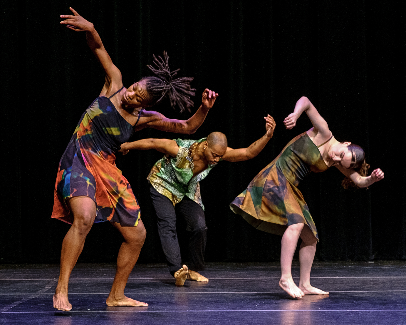 Three dancers, two women and a man in colorful tie dyed-like costumes, hair flying, with swinging arms bent over.