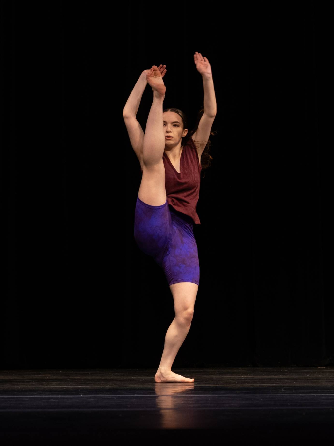 A woman dancer, dressed in shorts and sleeveless shirt in purple hues, battements her right leg and both arms as she looks to the audience.