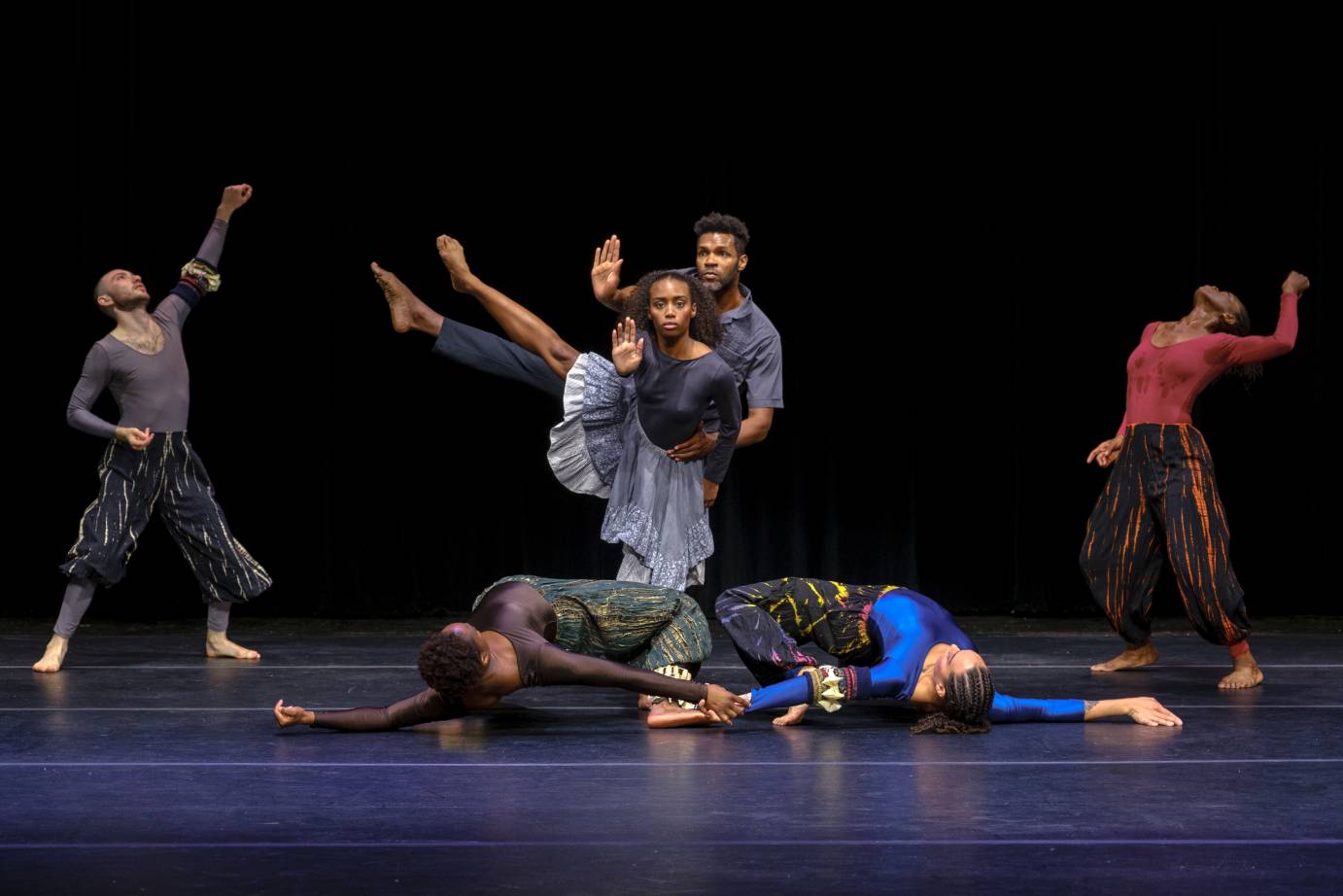 Two dancers rolling intertwined on the floor as a male and female duet with right legs extended stand behind them while two dancers on either side lift their fists and faces to the sky