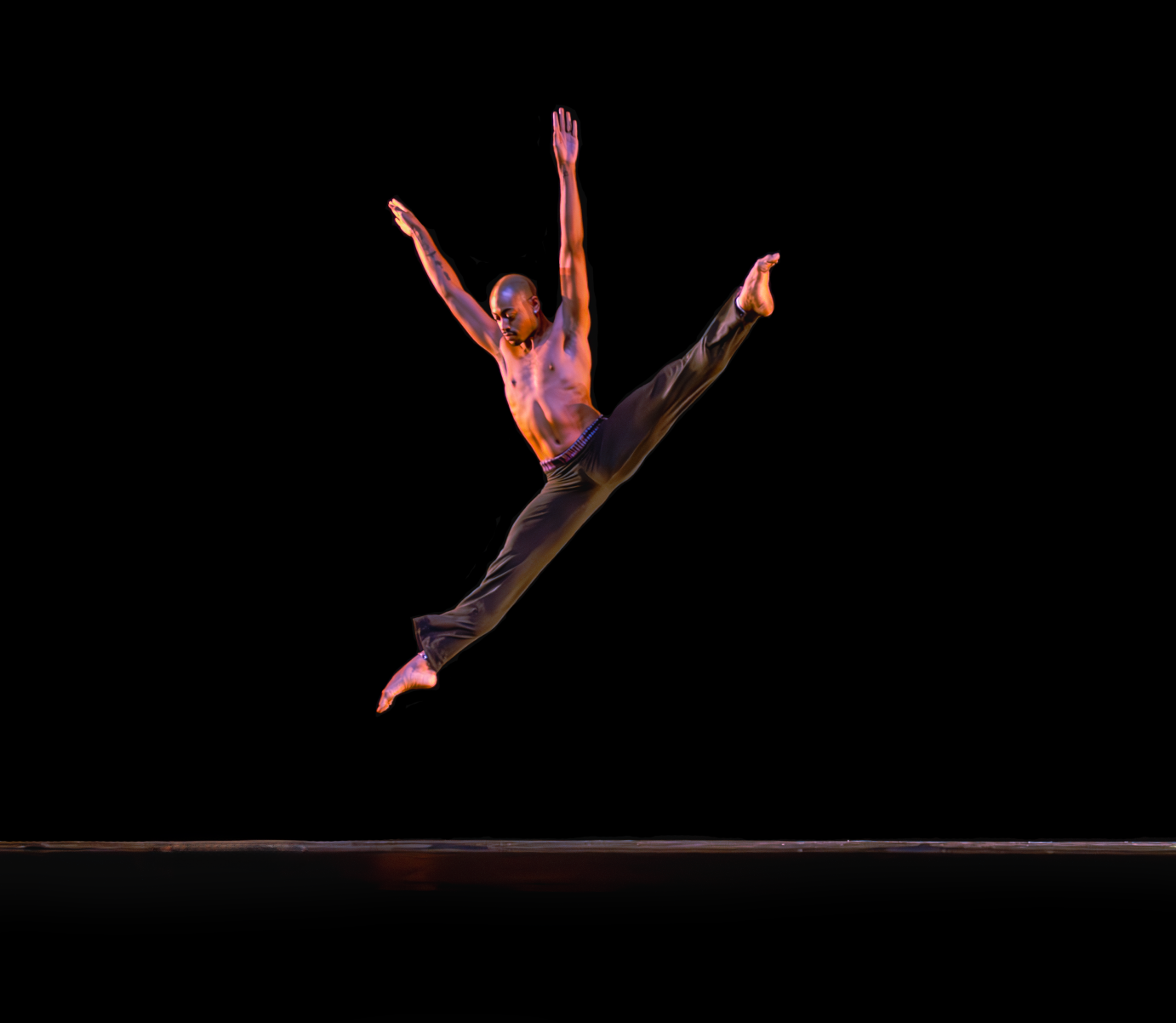 A man arms stretched on a diagonal, face looking down, jumps in a big center split leap.