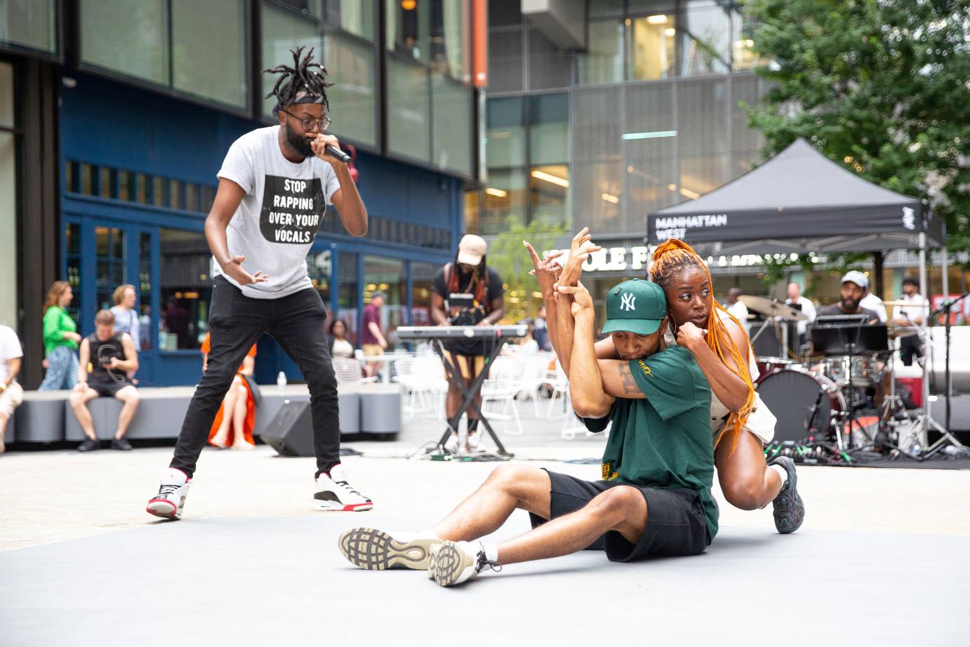 a rapping singer addresses two dancers one in green who is sitting on the ground and the other in white who clasps onto her partner's back their arms intertwined 