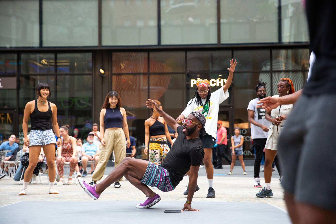 a bearded dancer in colorful print shorts and matching shoes balances on his hand and one foot in the middle of a floor series as his fellow dancers surround him cheering him on