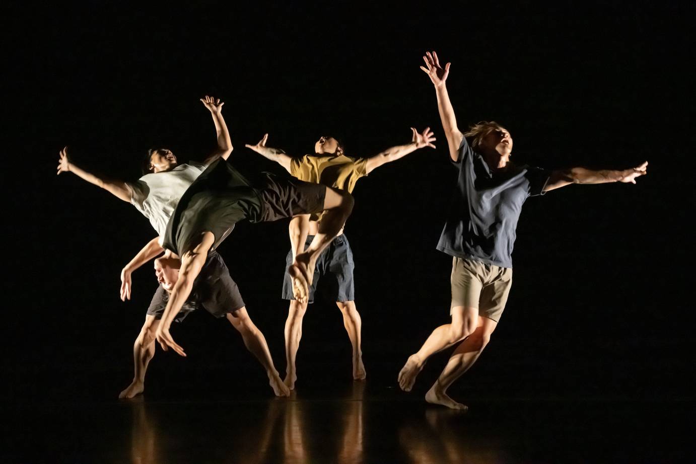 Three men jump in air with arms extended while a fourth man back flips and is caught on camera in midair. The men are dressed casually in shorts and shirts.