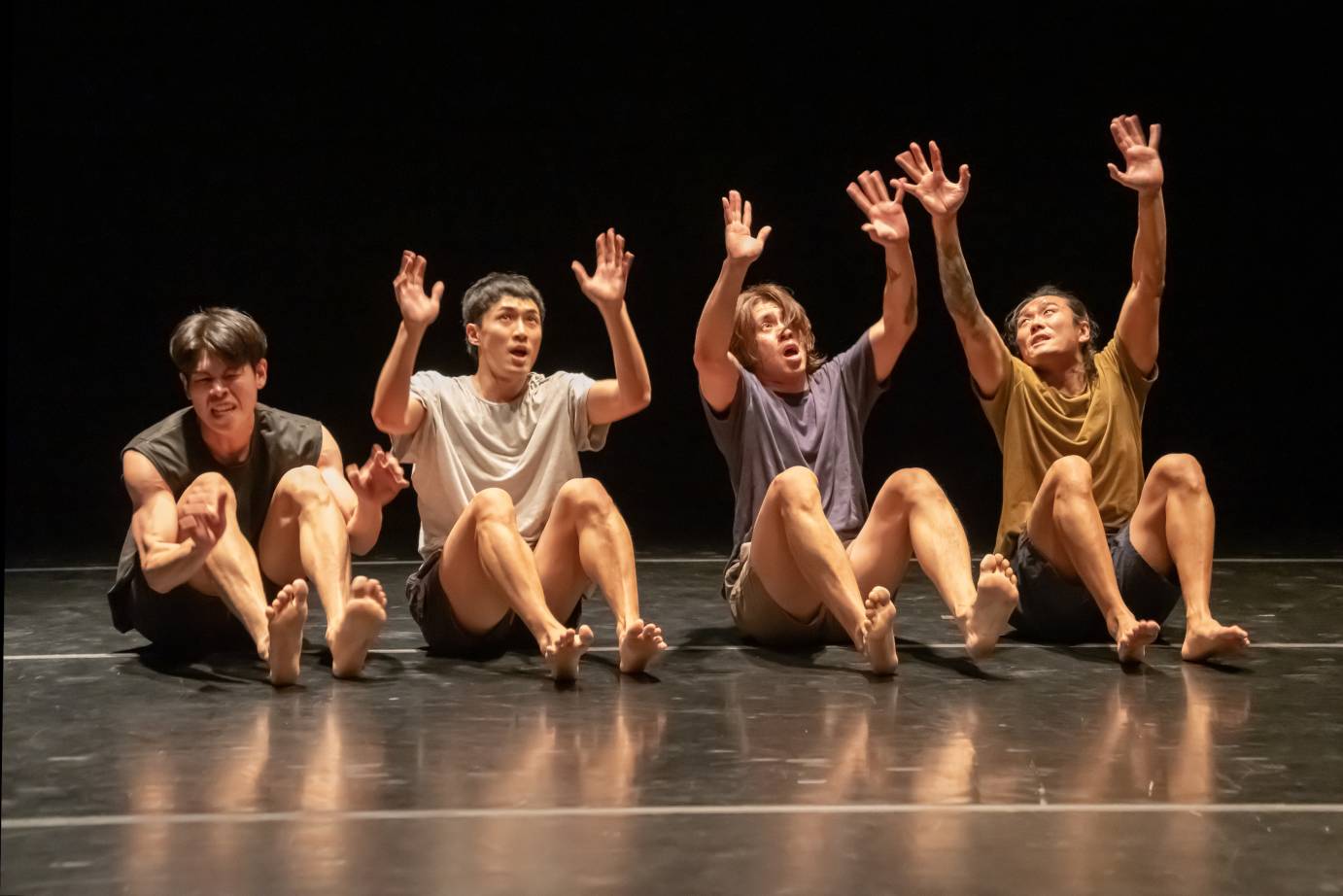 Four men sit side by side in unison with arms in the air and feet flexed.