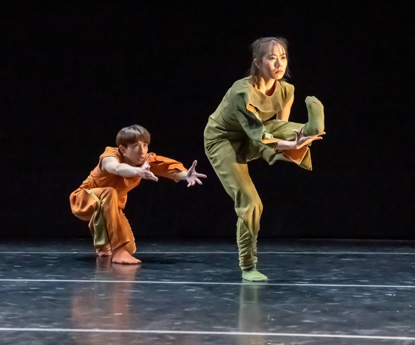 A woman dressed in a green top and pants stands on her right leg. Her left leg is extended, foot flexed with her fingers splayed holding her heel. She's wearing green socks and looking pensively on the diagonal. He, in orange, is crouched behind her with arms extended. His eyes looking up at her head.