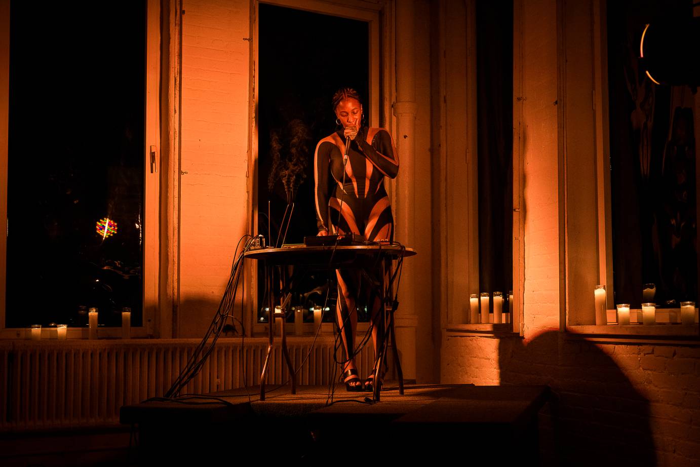 by windows in a loft lit by candles an artist in a unitard of black covered with revelatory triangles showing the artists skin, Renata Anakwe, appears to be DJing and singing simultaneously