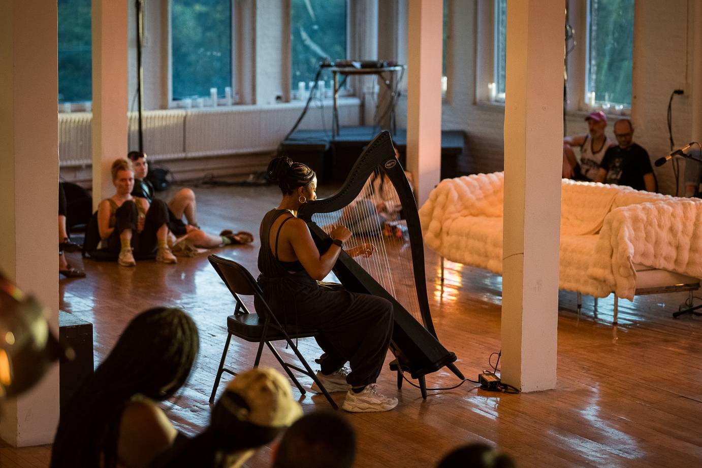 as audience members lean against pillars in a loft space, a black woman in a black jumpsuit with white sneakers place a harp, as she faces a white wooly sofa